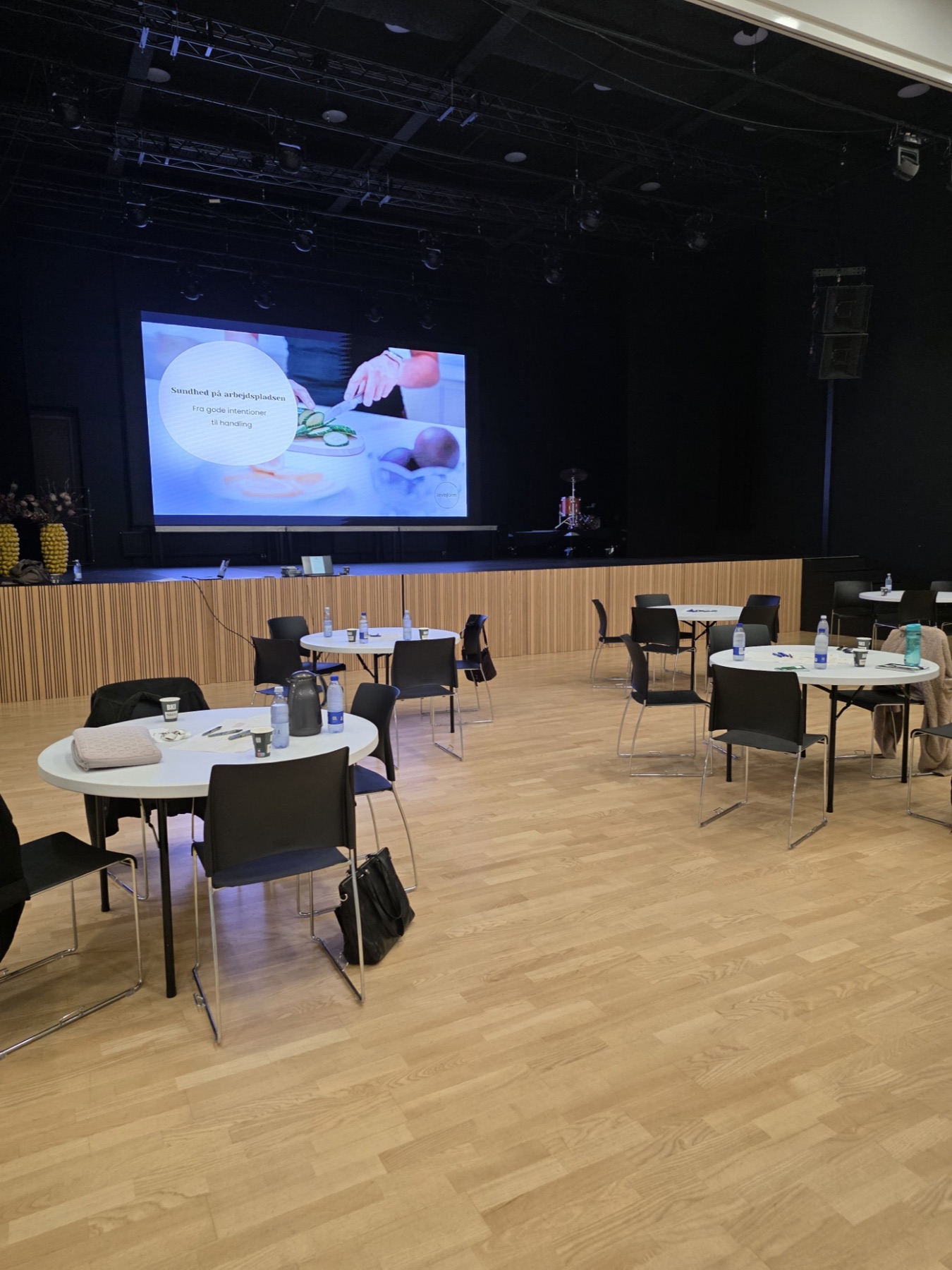 Lokale gjort klar til workshop med runde borde og skærm med titlen Sundhed på arbejdspladsen